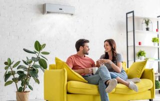 Couple relaxing on a yellow couch beneath an air conditioner, highlighting the best time of year to clean and maintain home air ducts.