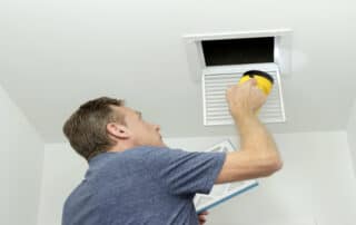 Man inspecting air ducts shining a flashlight through a small square ceiling vent into ducting pipes