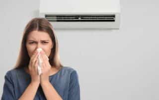 Woman sneezing near an air conditioner unit, illustrating how poor indoor air quality, mold, or unclean AC filters can circulate allergens and bacteria, potentially causing illness or allergy-like symptoms. Highlights the importance of regular air conditioner maintenance for healthier air.