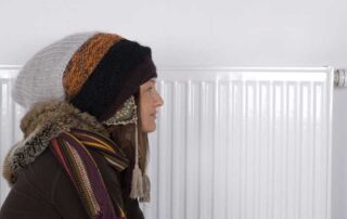ChatGPT said: Person dressed in warm winter clothing standing beside a home radiator, symbolizing energy-efficient heating solutions. Represents practical ways to stay warm and comfortable indoors while reducing winter heating bills through improved insulation and efficient heating systems.