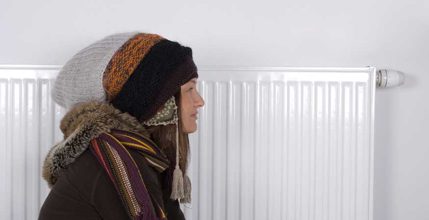 ChatGPT said: Person dressed in warm winter clothing standing beside a home radiator, symbolizing energy-efficient heating solutions. Represents practical ways to stay warm and comfortable indoors while reducing winter heating bills through improved insulation and efficient heating systems.